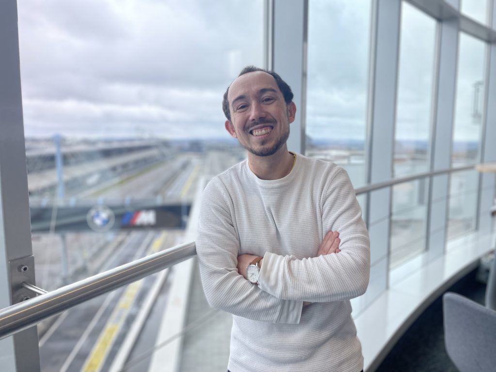 Mann mit dunklen Haaren und weißem Pullover schaut lächelnd in die Kamera.